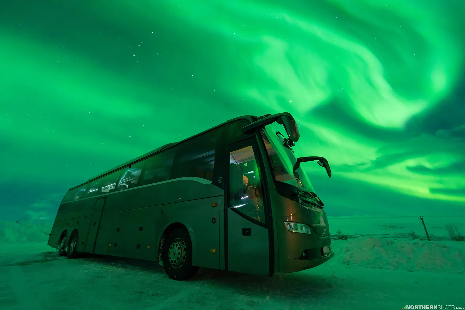 Exterior of the NorthernShots tour bus parked under a vibrant Northern Lights during an Arctic night.