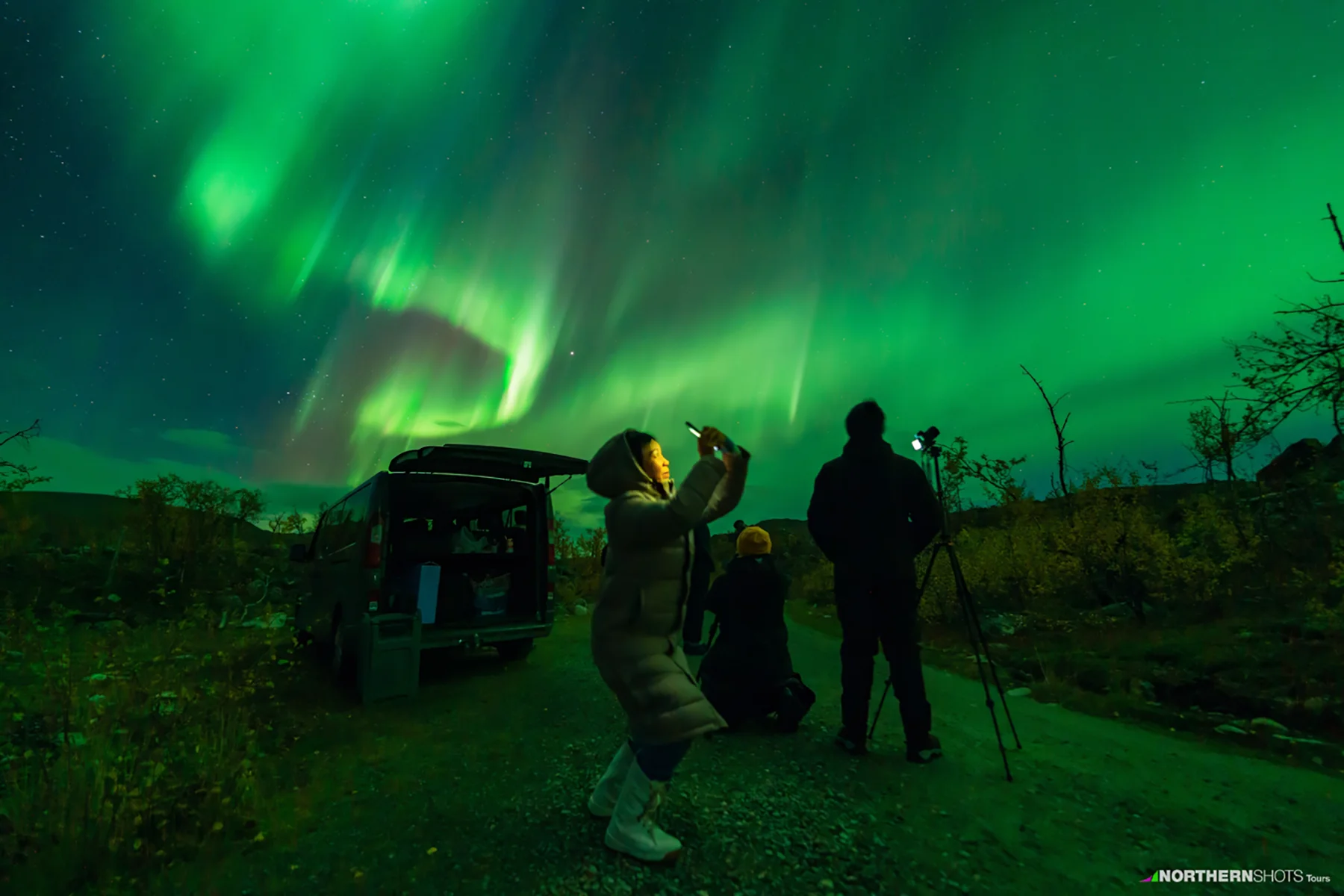 Travelers photographing the Northern Lights with tripods set up behind an open van on a gravel road.