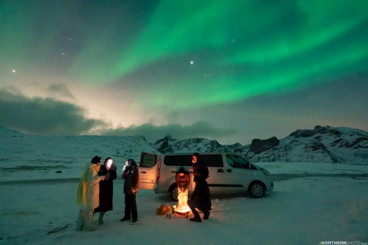 Guests gathered by a campfire in front of their van with a massive aurora display and moonlight above.