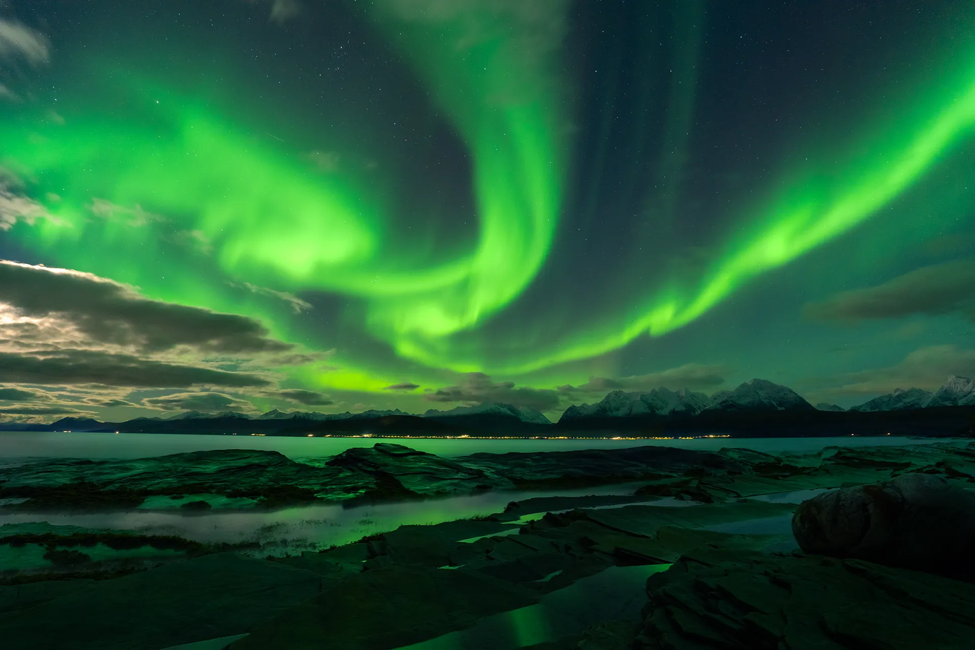 The Lyngen Alps at night viewed from a rocky shoreline, with vivid aurora visible through moonlit clouds.