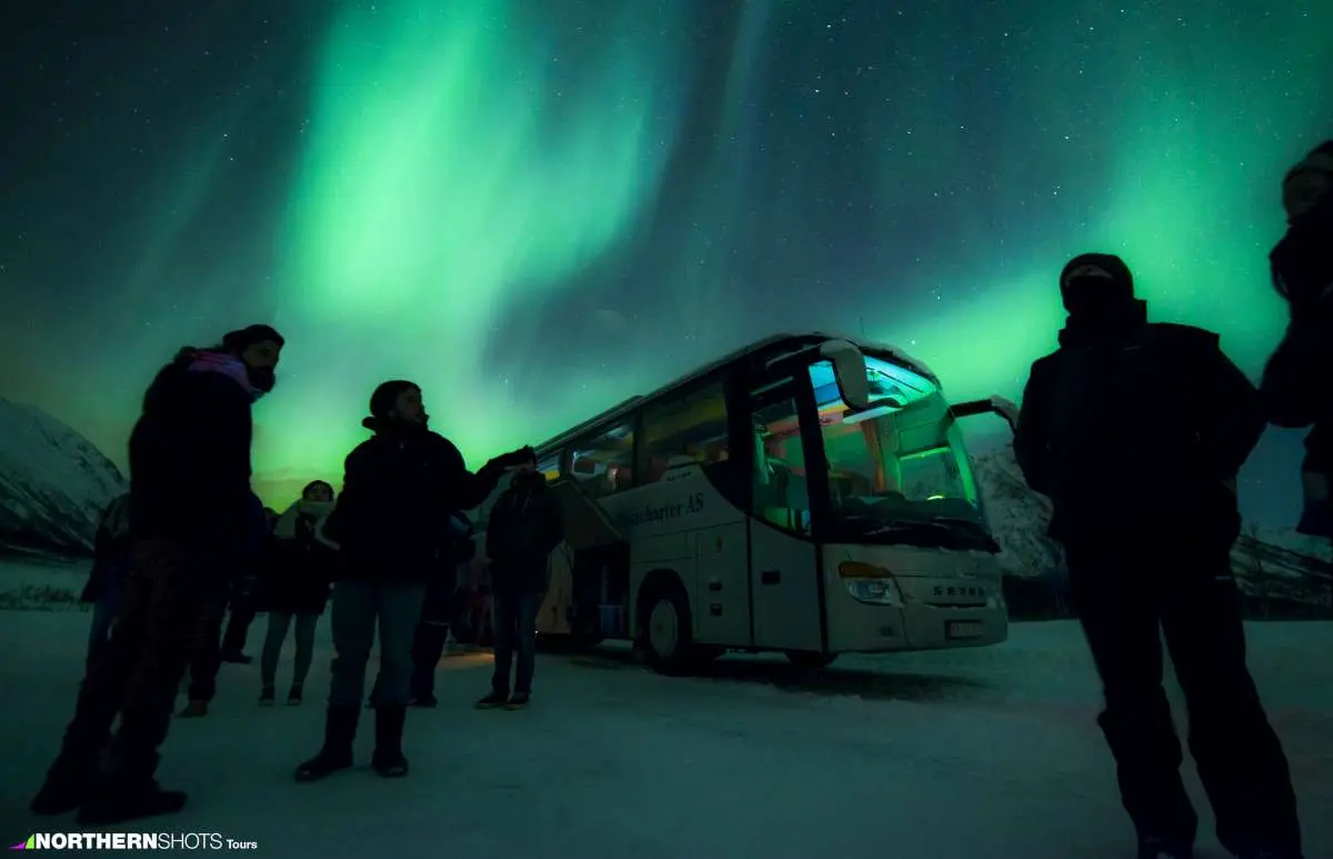 Silhouetted guests standing outside their bus, illuminated by a vivid green Northern Lights display overhead.