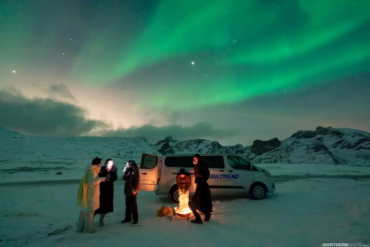 Guests gathered by a campfire in front of their van with a massive aurora display and moonlight above.