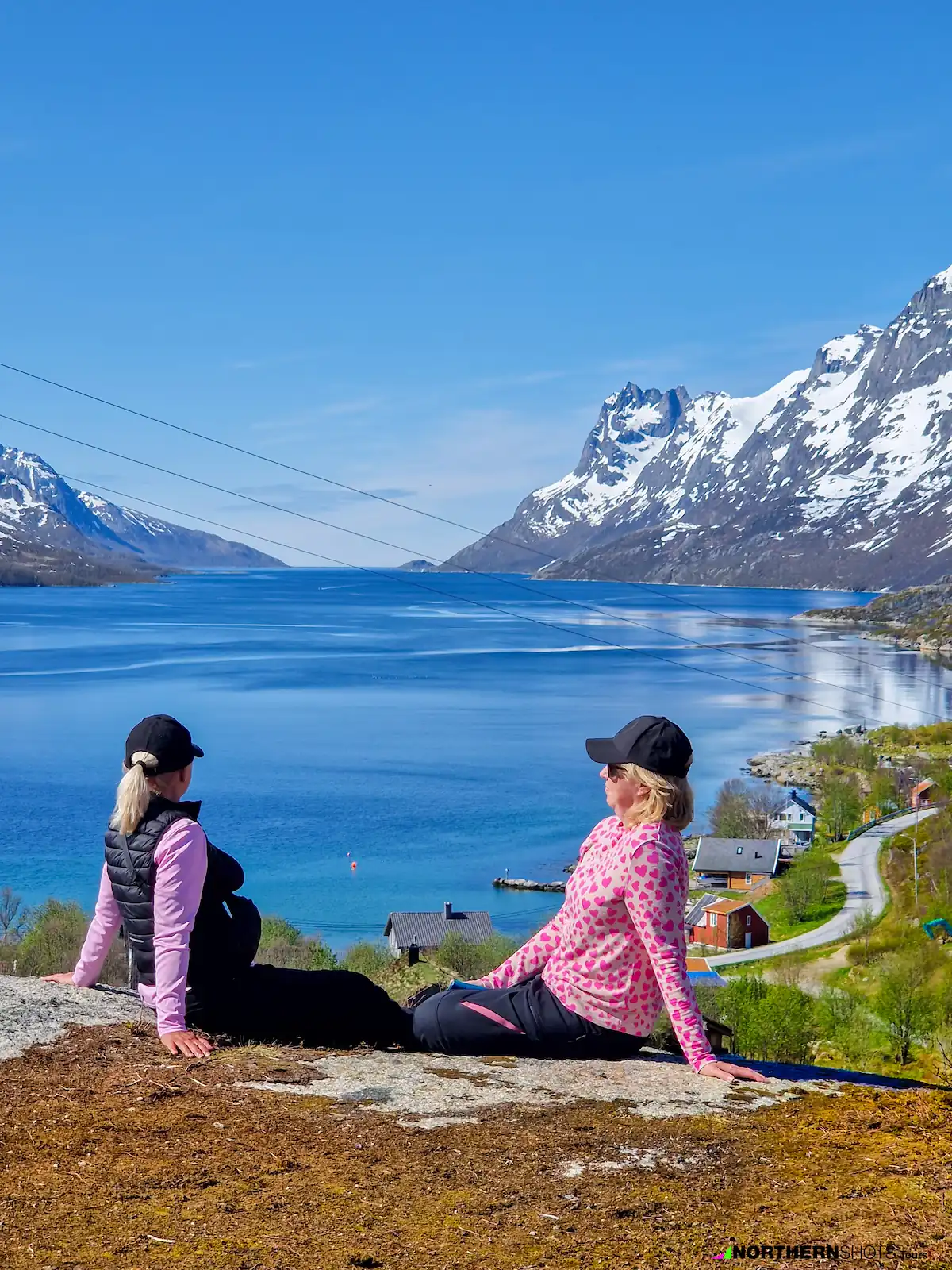 Tourists relaxing on mountain top near Tromsø, overlooking fjord with snowy peaks in summer.